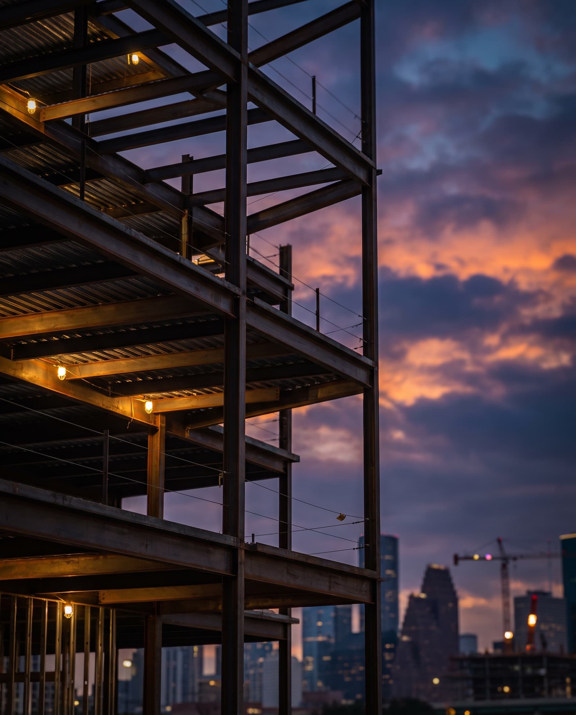 A mid-rise commercial steel-frame building under construction at dusk, low angle toward the sunset